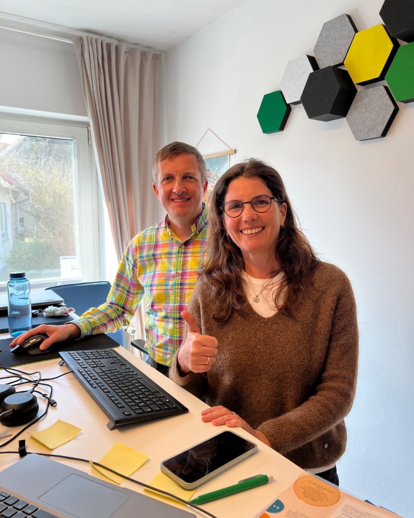 Kerstin und Marc (Geschäftsführung) stehen nebeneinander am Schreibtisch und lächeln in die Kamera.