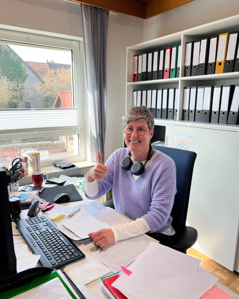 Mitarbeiterin Simone zeigt lächelnd den Daumen in die Kamera. Sie sitzt an ihrem Schreibtisch vor einem Ordnerregal.