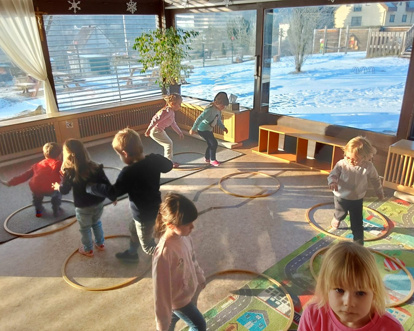 Kinder hüpfen im Kreis von Ring zu Ring im Gebäude des Kindergarten Lengfurt. Durch die große Fensterfront sieht man den schneebedeckten Garten.