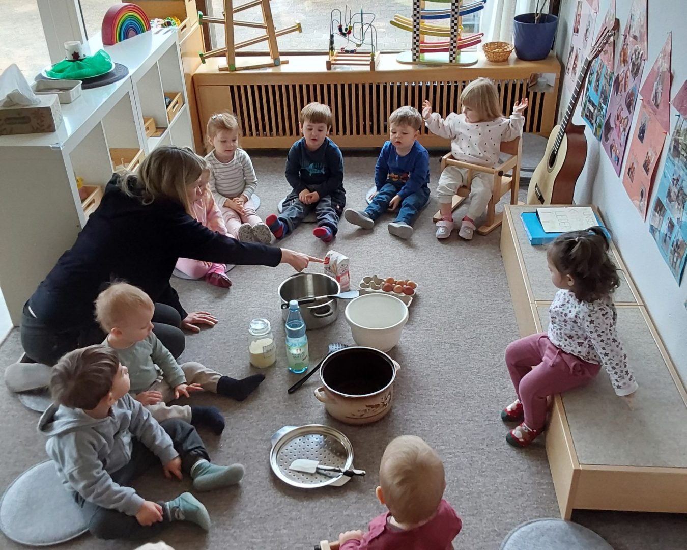 Kinder sitzen in einem Kreis um alle Zutaten, die man zum Backen braucht. Eine Erzieherin erklärt alles.